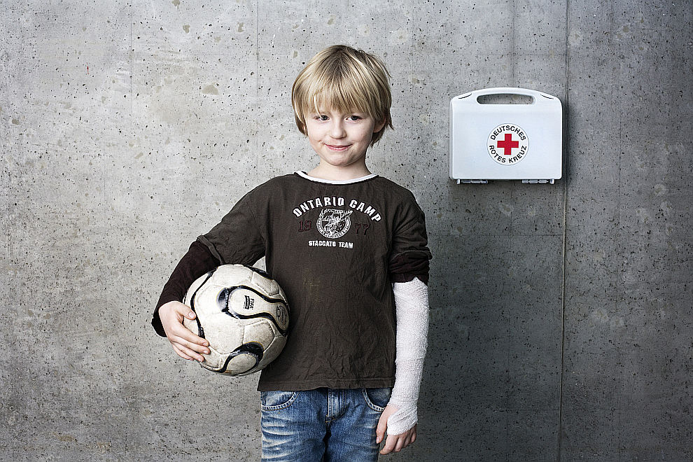 Junge mit verbundenem Arm hält einen Ball. Neben ihm ein Verbandskasten mit dem Logo des DRK