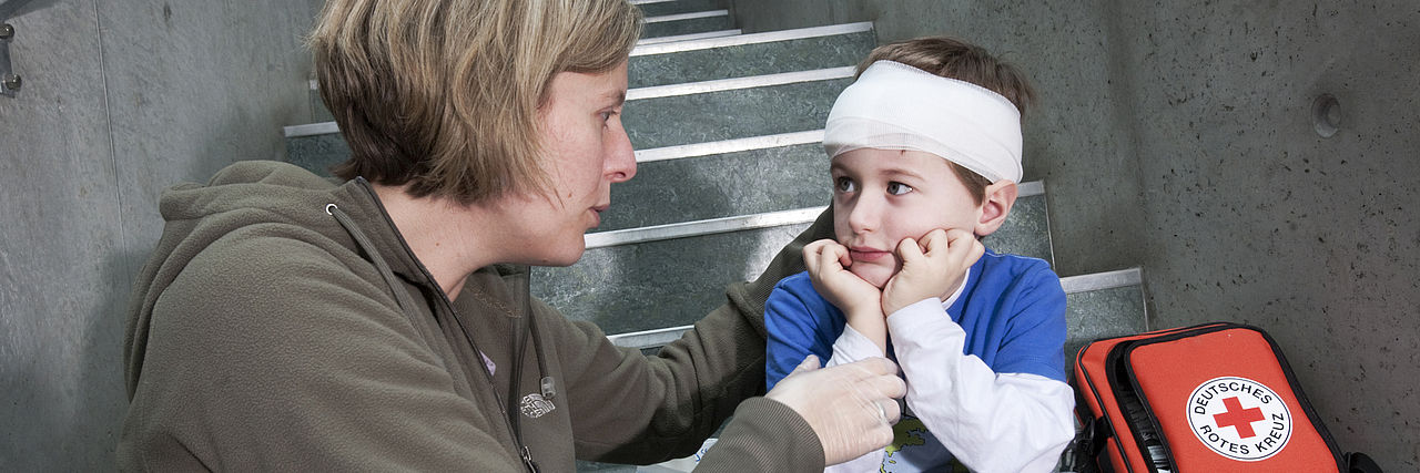 Mutter mit ihrem verletzten Sohn, er trägt einen Verband am Kopf. Neben dem Kind ein Verbandset mit dem Logo des DRK